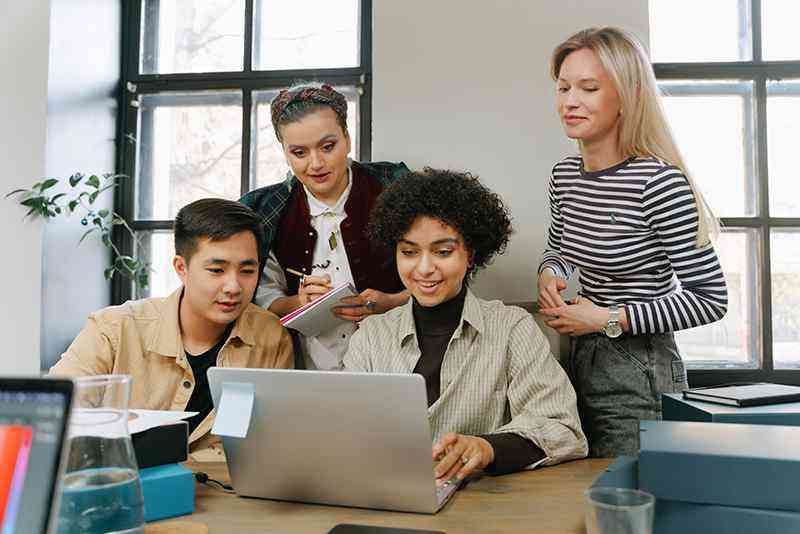 Four diverse team members gather around a laptop in a bright office, reviewing work together while one takes notes.