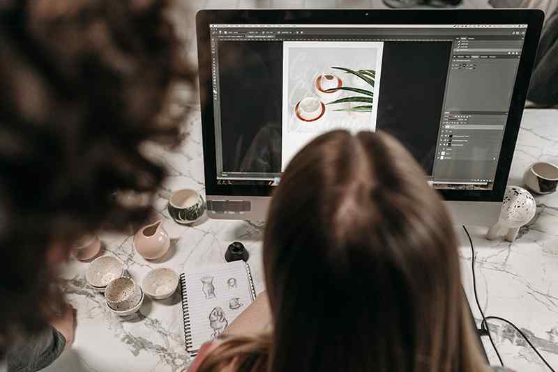 A designer works on digital artwork at a desktop monitor, surrounded by handmade ceramic pieces and design sketches on a marble table.