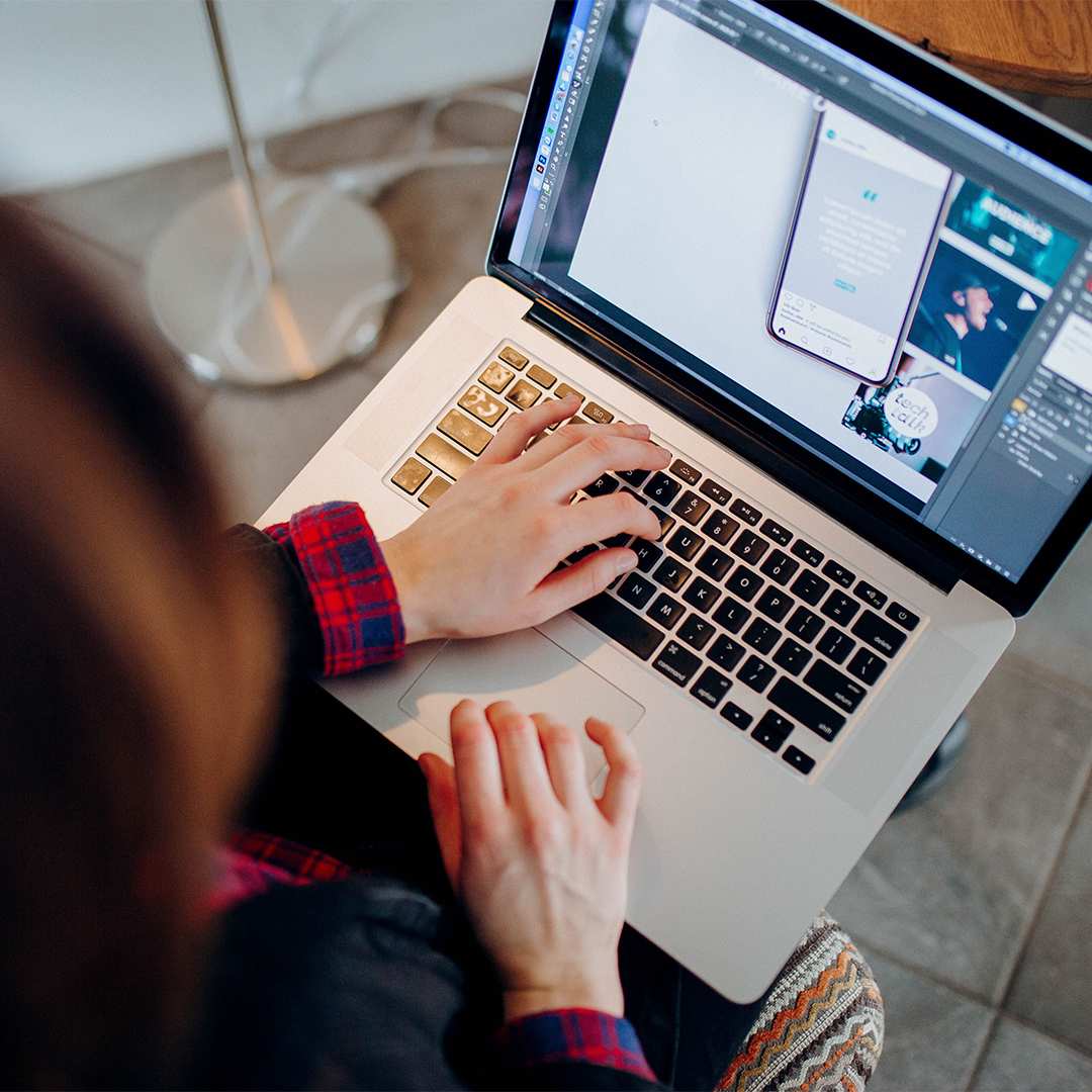 Overhead view of a person in a plaid shirt typing on a silver laptop, with mobile app and web design layouts visible on the screen.