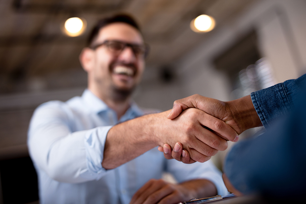 Smiling man in a light blue shirt shaking hands with another person, symbolizing a successful business agreement or new partnership in a warm office setting.