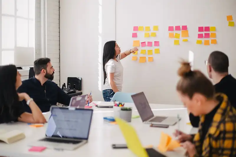 A woman pointing to colored sticky notes on a wall while colleagues with laptops look on during a brainstorming session