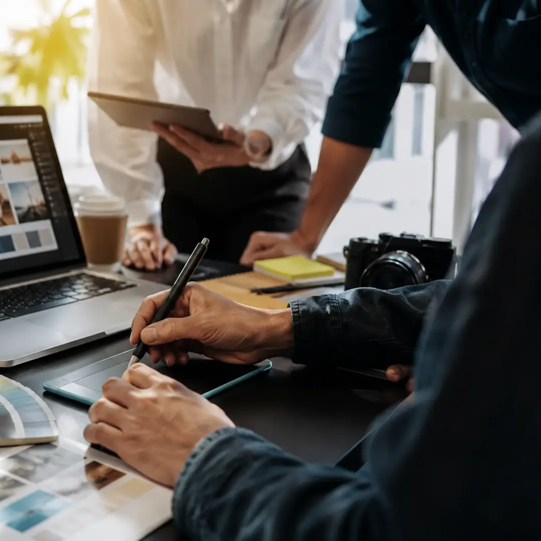 A graphic design team collaborating around a laptop and drawing tablet, reviewing branding concepts, design layouts, and creative assets in a modern office workspace.