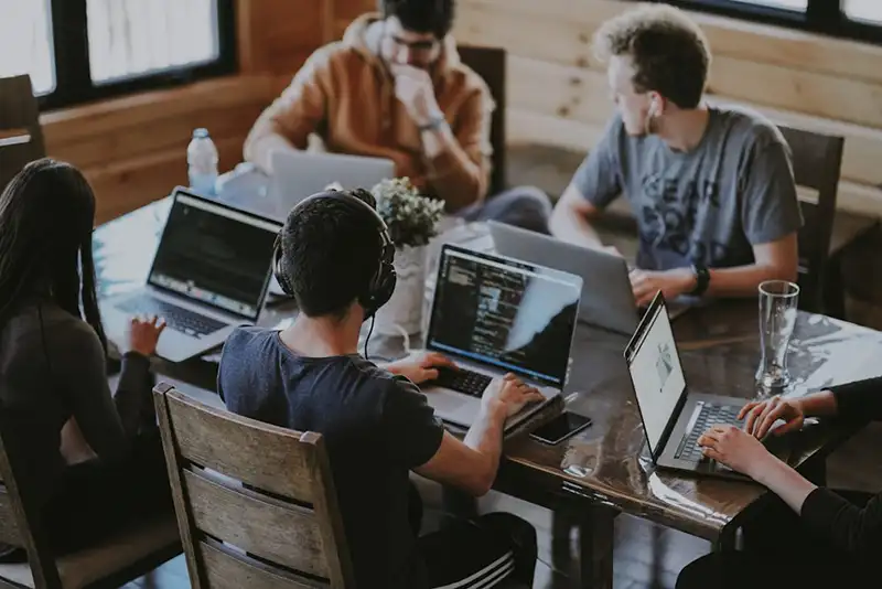 A group of colleagues wearing headphones and working on laptops together at a wooden table in a casual workspace