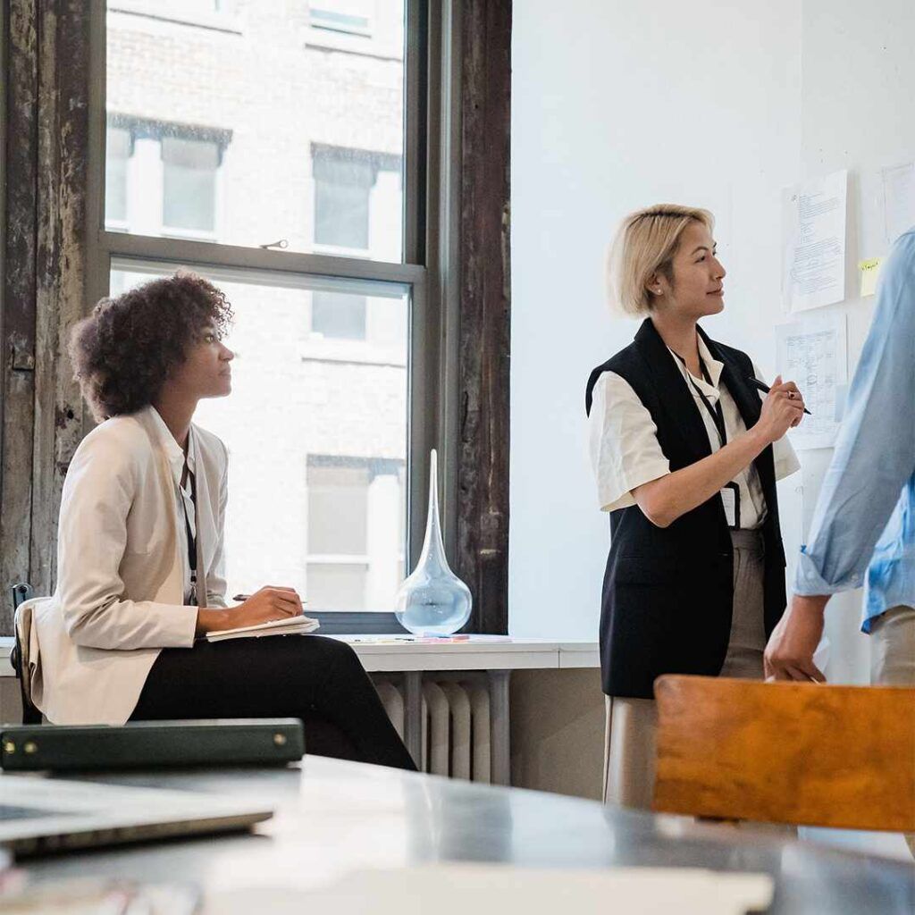 Two women collaborate in a bright industrial-style office: one stands explaining ideas near pinned documents, while the other sits taking notes, with natural light streaming through a large window.