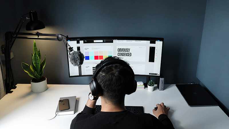 Back view of a person wearing headphones working at a white desk with a large curved monitor, studio microphone, desk lamp, snake plant, smartphone, and laptop on a dark blue wall background