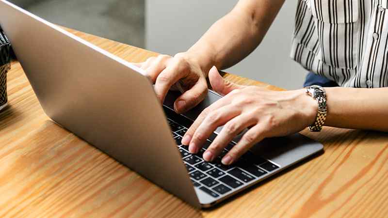 Close-up of a person in a striped shirt typing on a sleek silver laptop placed on a light wooden table, wearing a silver wristwatch, in a bright workspace.