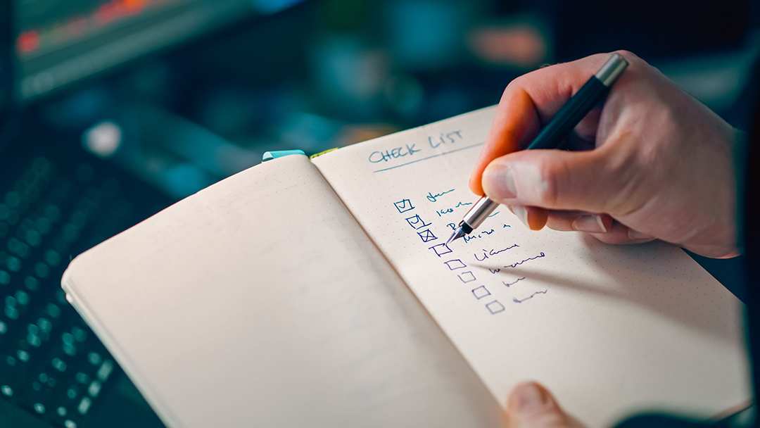 Hand writing a checklist in a dotted notebook with a pen, laptop keyboard blurred in background, planning a brand audit step-by-step