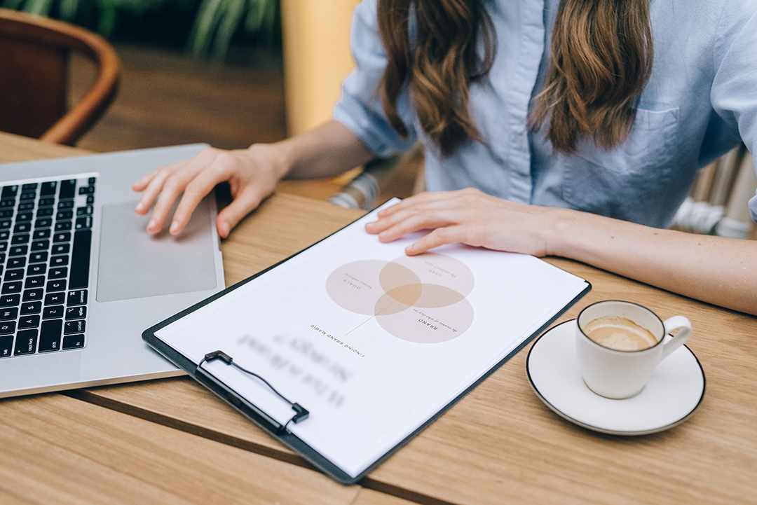 Woman reviewing a brand strategy planning sheet with Venn diagram on a clipboard, silver laptop and latte cup on light wooden table, soft natural lighting, professional branding and design work concept