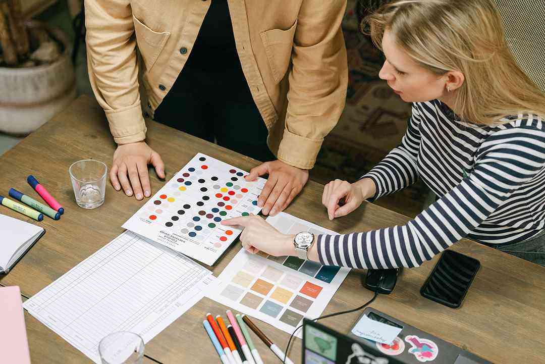 Two designers reviewing and comparing brand color palettes and visual identity charts on a wooden worktable during a brand audit, with markers, swatch sheets, and design tools spread across the desk