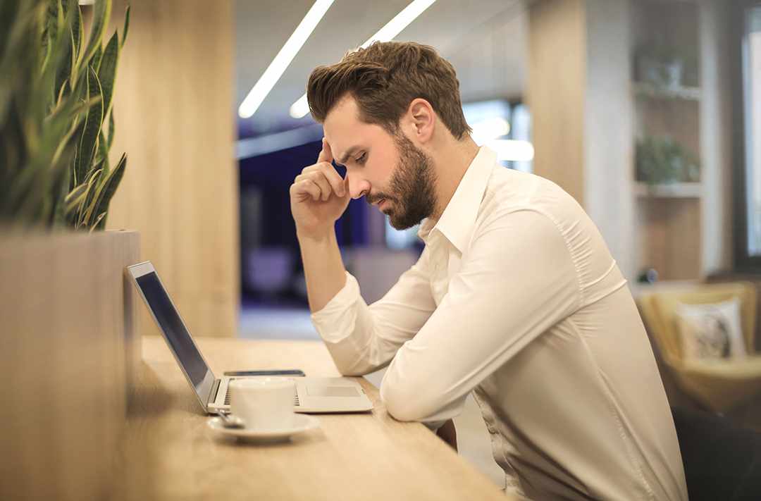 Professional man in a white shirt sits at a desk with a laptop, resting his hand on his forehead with a thoughtful, stressed expression in a modern office interior
