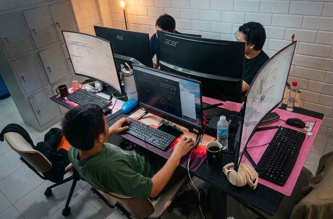 Three team members working at a shared desk with multiple curved monitors, designing branding and reviewing project data at Pugo Design Studio office