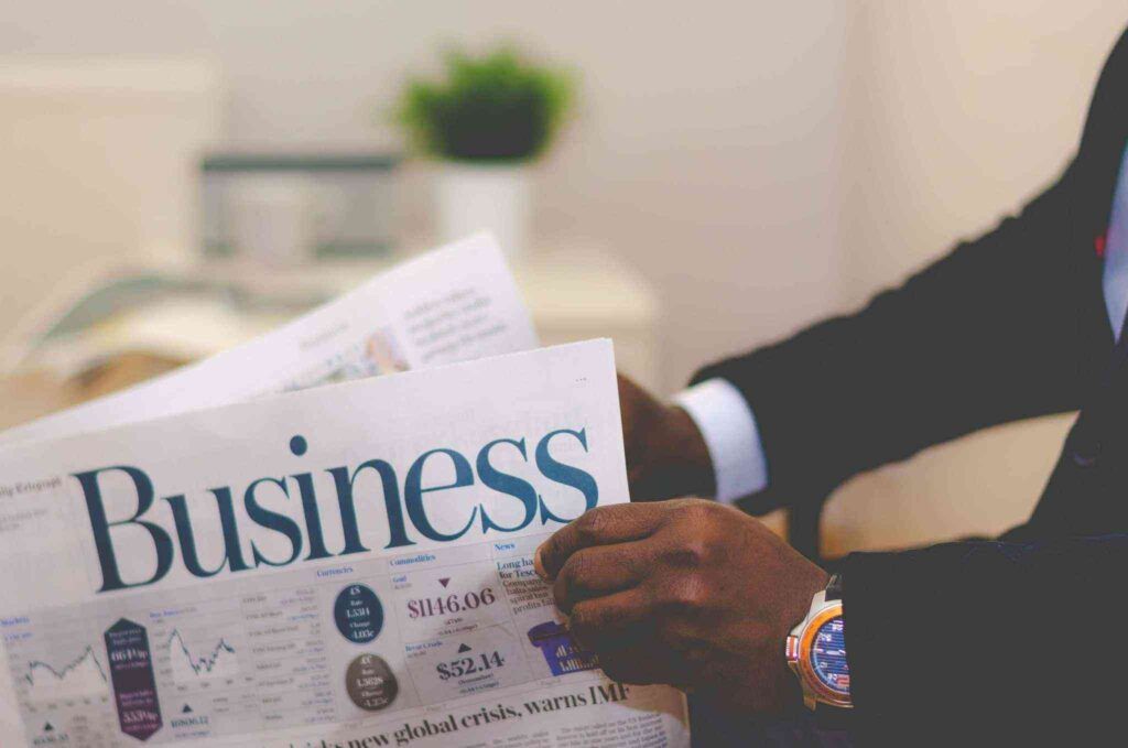 Close-up of a professional in a dark business suit holding a newspaper prominently displaying the word "Business," with financial charts, market data, and currency figures visible on the page; blurred office background with a small potted plant.
