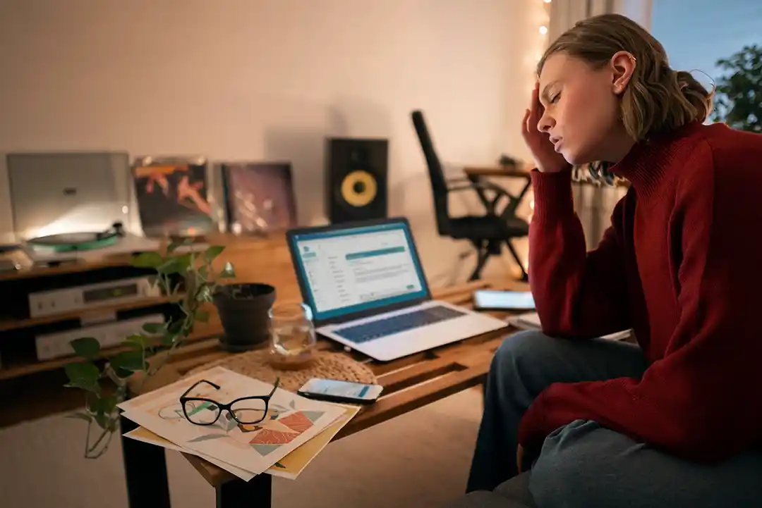 Overworked creative professional sitting at a desk with laptop and design materials, holding head in hand while experiencing burnout and project bottleneck pressure