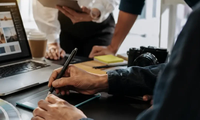 A graphic design team collaborating around a laptop and drawing tablet, reviewing branding concepts, design layouts, and creative assets in a modern office workspace.