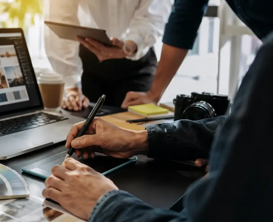A graphic design team collaborating around a laptop and drawing tablet, reviewing branding concepts, design layouts, and creative assets in a modern office workspace.